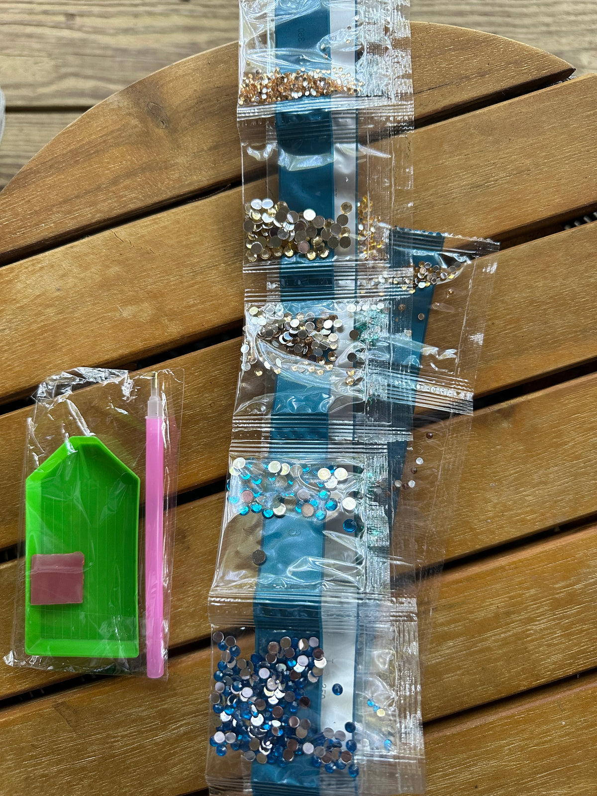 A DIY craft kit laid out on a wooden slatted surface, including a clear plastic organizer filled with blue, silver, and gold rhinestones, a green diamond-picking tool, and a pink-handled tool in a clear plastic bag.
